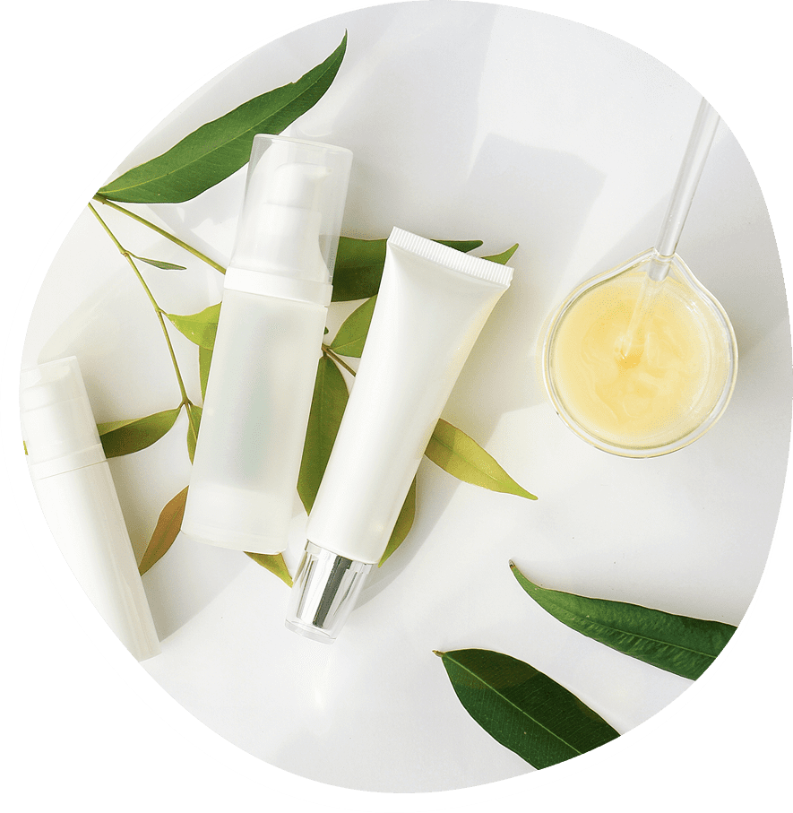 Skincare products and a glass bowl with cream on a white surface, surrounded by green leaves.