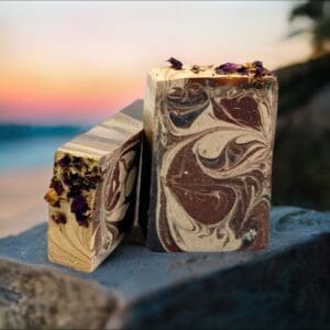 Two handcrafted Black Amber & Lavender soaps featuring brown and cream swirls, topped with dried flowers, rest on rocks against a blurred sunset background by the water.