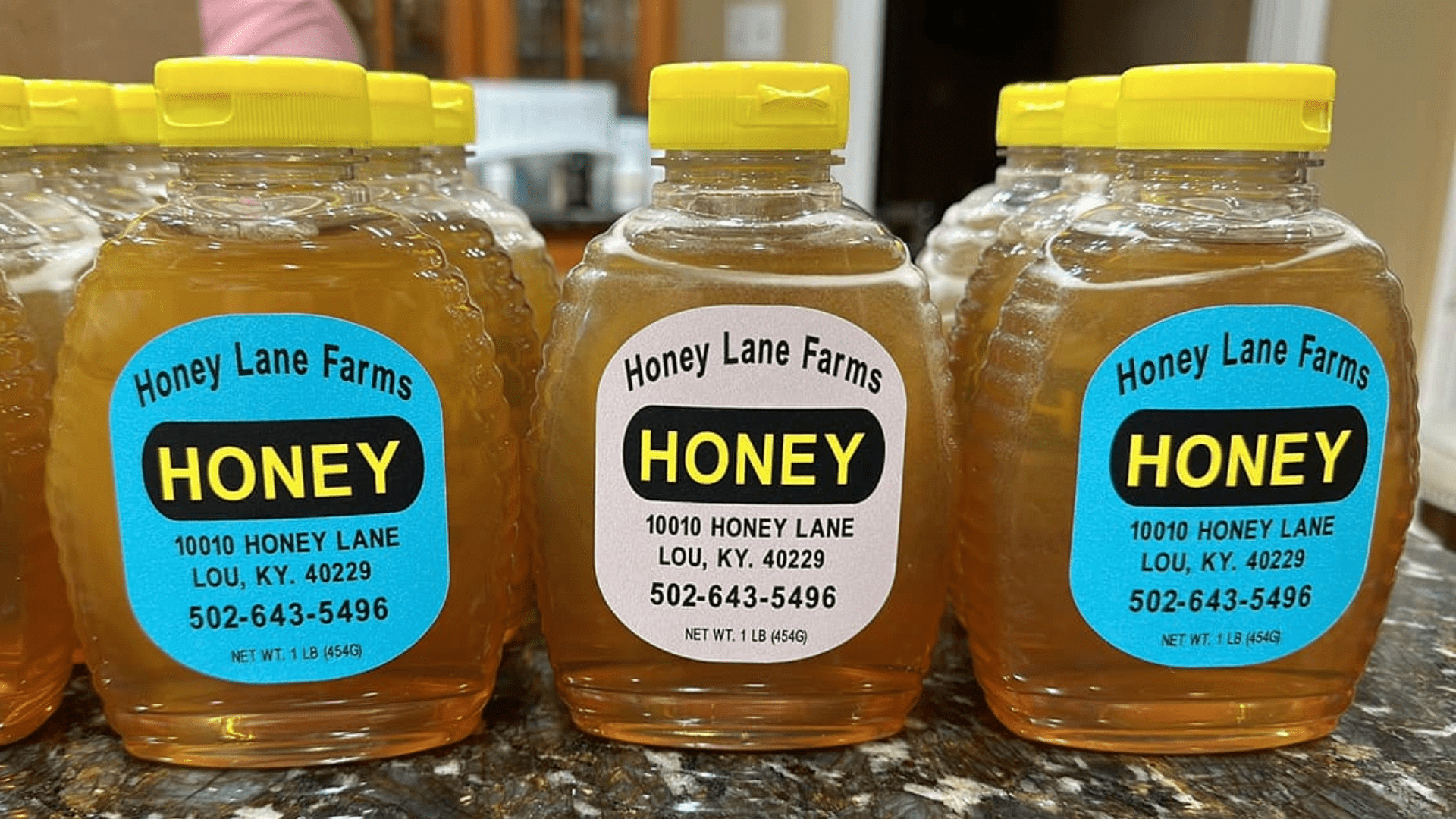 Three plastic bottles of Honey by Honey Lane Farms with yellow lids, displaying labels with the farm's name, address, and contact number on a countertop.
