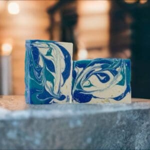 Two bars of Barber Shop handmade soap featuring blue, white, and teal swirls rest on a stone surface with a blurred background.