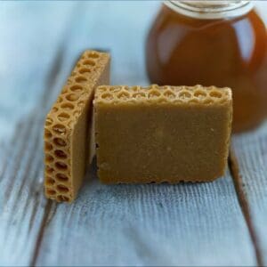 Two Honey Oatmeal soap bars with a honeycomb pattern rest on a wooden surface, accompanied by a jar of honey in the background.