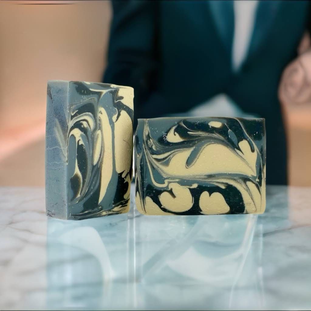 Two "Suit & Tie" soap bars with black and gold marbled patterns rest on a glossy surface, with a blurred person in a suit in the background.