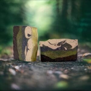 Two "Hike Your Own Hike" artisanal soaps, featuring green, brown, and cream swirls, rest on a forest-like surface with a blurred background.