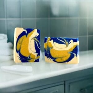 Two bars of Crisp Cotton soap with swirling yellow, blue, and white patterns sit on a bathroom counter near folded towels and a soap dish.