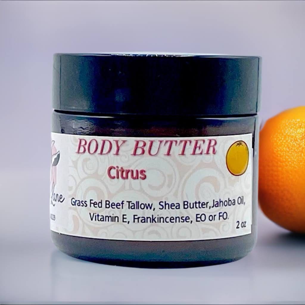 A jar of Citrus Body Butter - 2oz sits with an orange, labeled as featuring grass-fed beef tallow, shea butter, jojoba oil, and vitamin E.