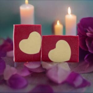 Two Love Spell heart-themed red soaps featuring white heart designs, surrounded by rose petals and lit candles.